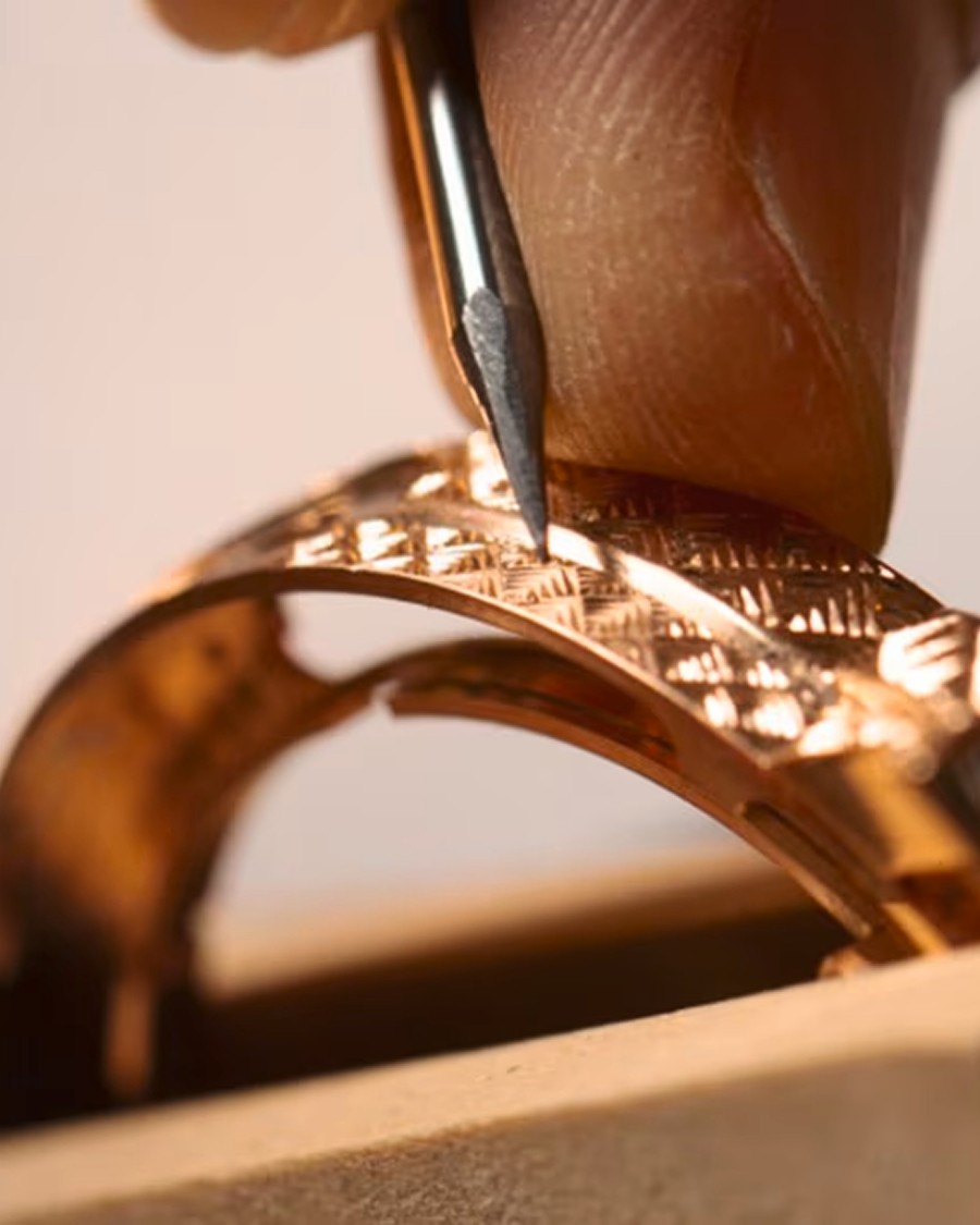 Detailed view of a hand engraving a decorative pattern onto a golden watch case using a specialized tool.