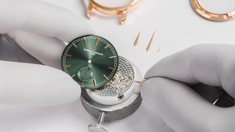 Two hands in white gloves are setting up a watch with a green dial with gold details