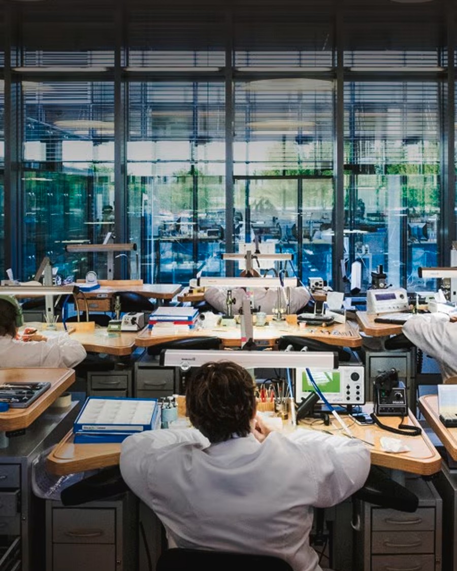 Wide view of a watchmaking workshop with artisans working at desks in a modern, glass-walled space.