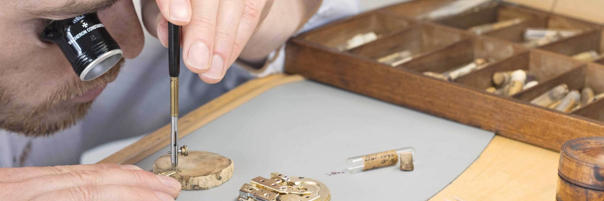 Watchmaker assembling watch parts using a screwdriver at a wooden workbench with organized tools.