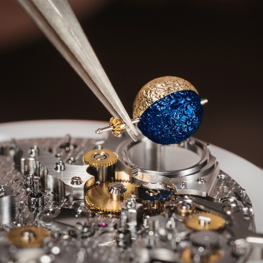 Watchmaker assembling a round, gold and blue element onto a watch movement with tweezers.