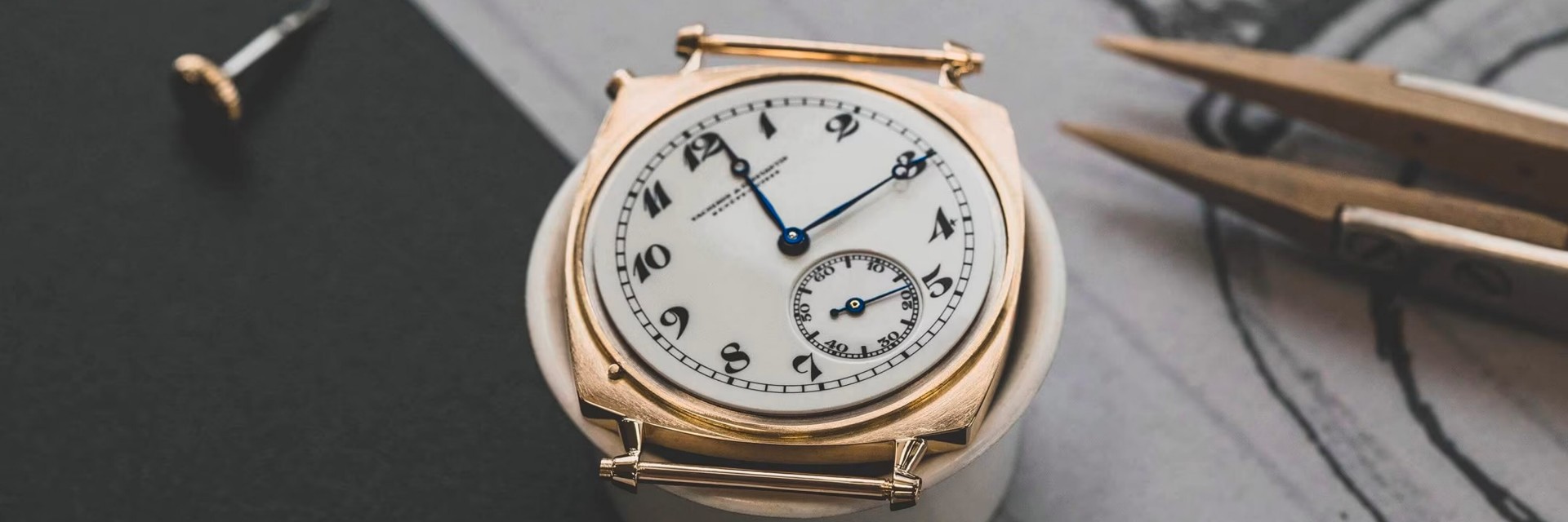 Vacheron Constantin watch with a white dial, black numerals, and gold case, placed on a table with tools in the background.