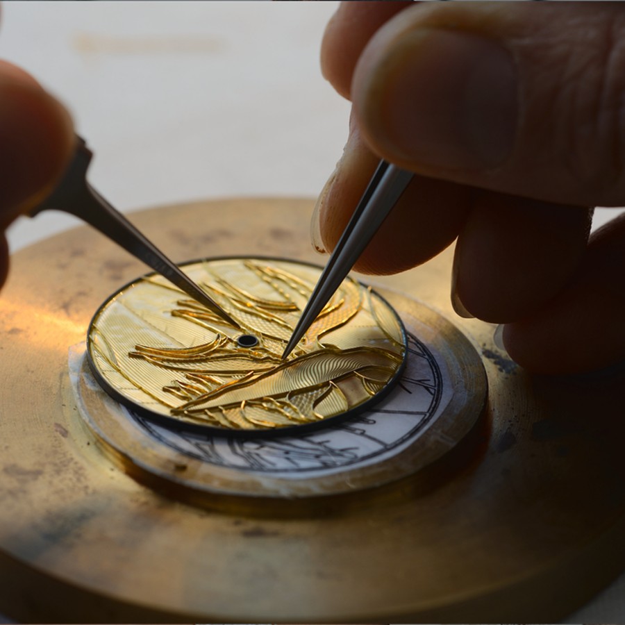 Hands working on an engraved golden watch dial using fine tweezers and tools.